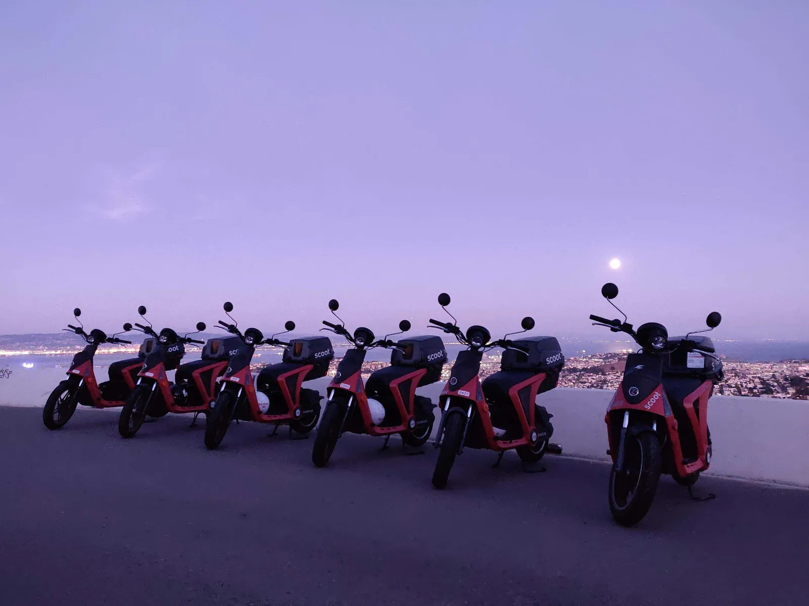 Row of red Scoot electric scooters parked on a hilltop overlooking a city at sunset