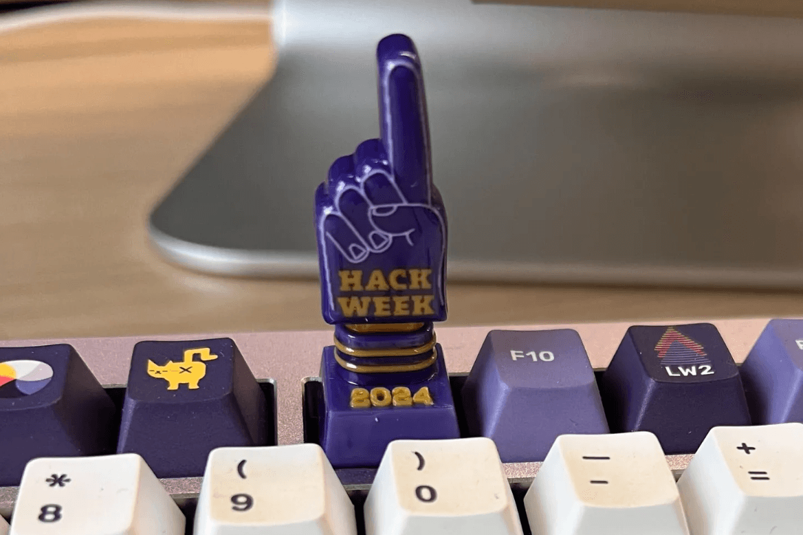 A picture of a keycap for a mechanical keyboard that is on the F9 key. The keycap has a "foam finger" in purple that says "Hack Week 2024"